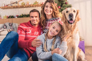 gülümseyen aile ile Noel ağacının yanında hediyelerle oturan golden retriever köpek
