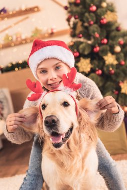 Noel Baba şapkası ve having fun evde Noel ağacı yakınındaki geyik boynuzları ile köpek mutlu çocuk