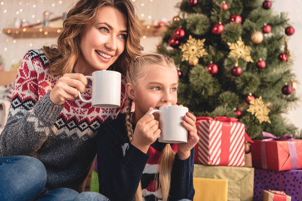 счастливая мать и дочь пьют какао возле рождественской елки дома
