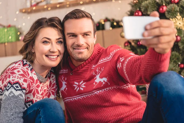 夫と妻の家にクリスマス ツリー近くのスマート フォンと Selfie を取ってのセーターで笑みを浮かべてください — ストック写真