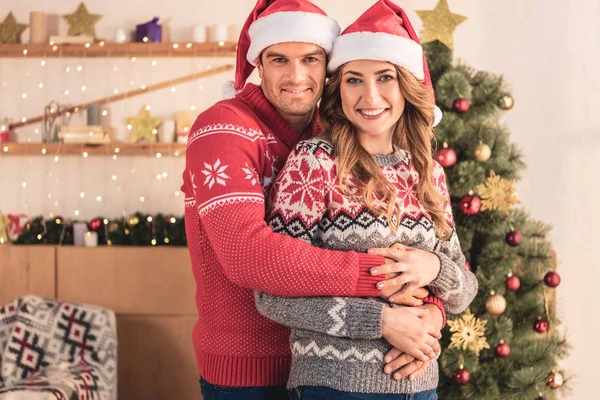 サンタ帽子ハグ妻近く自宅のクリスマス ツリーとカメラ目線で笑顔の夫  — 無料ストックフォト