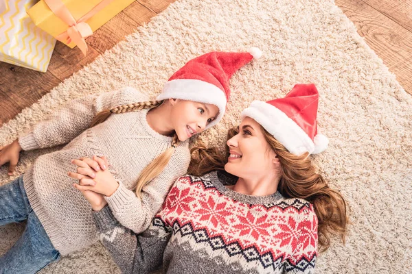 圣诞老人帽子上的母亲和女儿躺在地毯上 看着对方在家里的顶部视图 — 图库照片