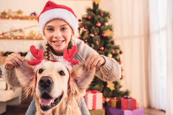 smiling kid in santa hat and golden retriever dog with deer horns having fun at home near christmas tree