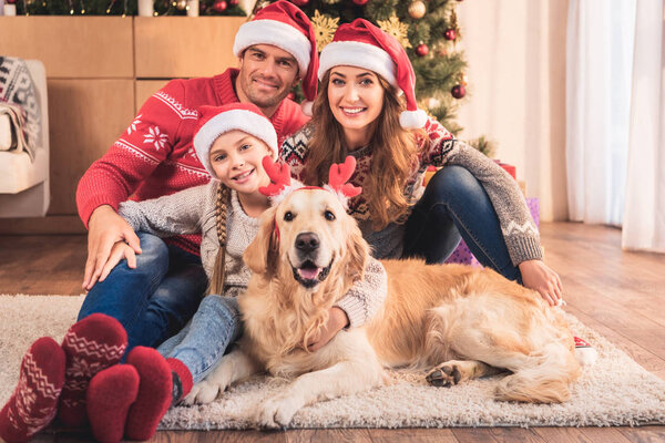 счастливая семья в шляпах Санта-Клауса с собакой в оленьих рогах, сидящей возле рождественской елки
