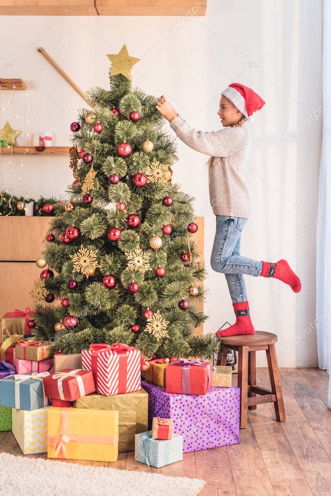 happy child in santa hat standing on stool and decorating christmas tree with gift boxes