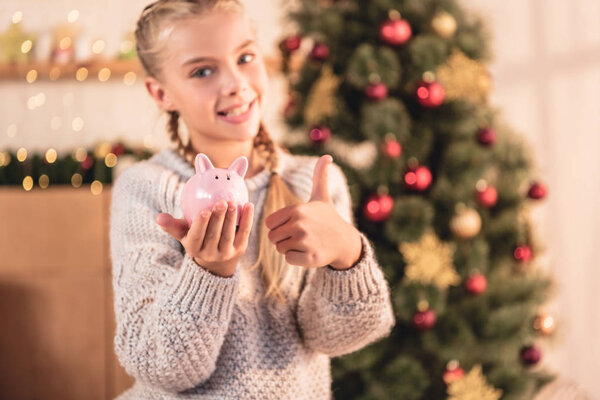 adorable child holding piggy bank and showing thumb up at home with christmas tree