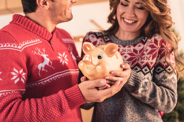 happy couple in christmas sweaters holding big piggy bank 