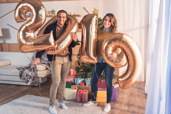 happy wife and husband holding 2019 new year golden balloons at home with christmas tree and presents