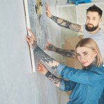 Young tattooed couple hanging painting on wall at new apartment