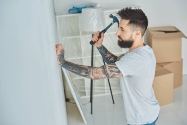 Yeni evde duvara çivi çekiç sakallı genç adam yan görünüm