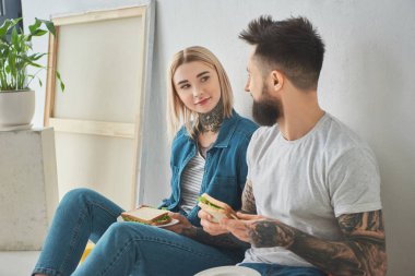 sandviç tutarak ve her diğer kat yeni evde otururken gülümseyen mutlu genç çift 