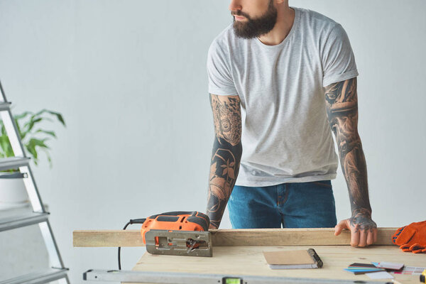 cropped shot of bearded man leaning at wooden table with electric jigsaw during home repair
