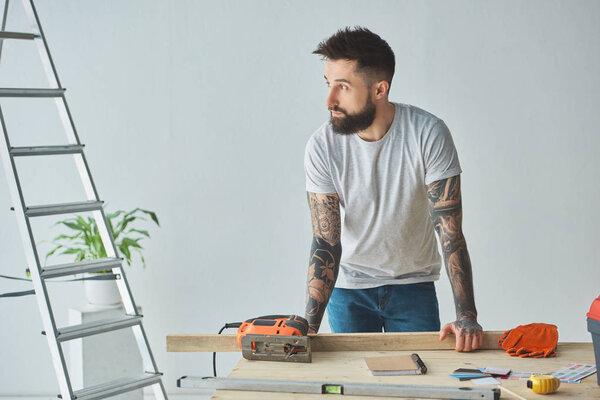 красивый бородатый мужчина склоняется за деревянным столом и смотрит в сторону во время ремонта дома
 