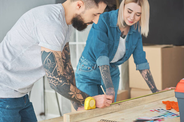 smiling young tattooed couple working with wooden plank and measuring tape 