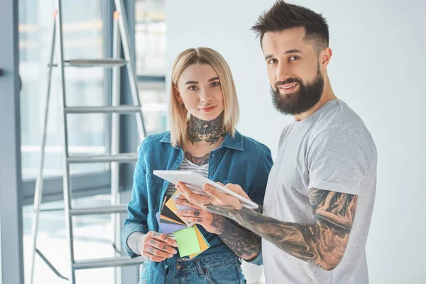 young tattooed couple using digital tablet and smiling at camera in new home