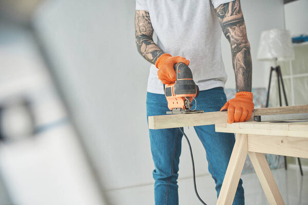 cropped shot of young tattooed man using electric jigsaw during home improvement