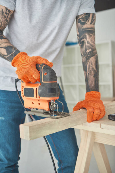 cropped shot of young tattooed man using electric jigsaw  