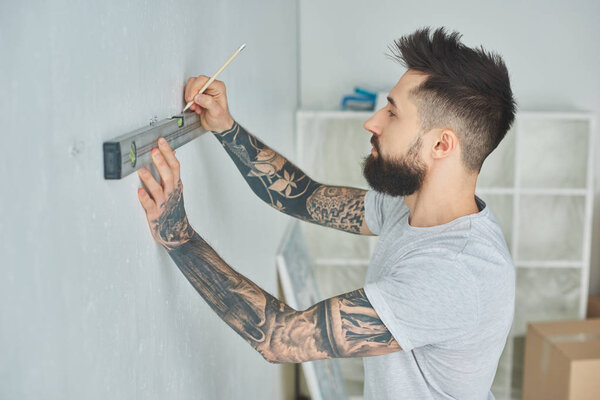 side view of tattooed young man holding level tool and marking wall with pencil during repairs