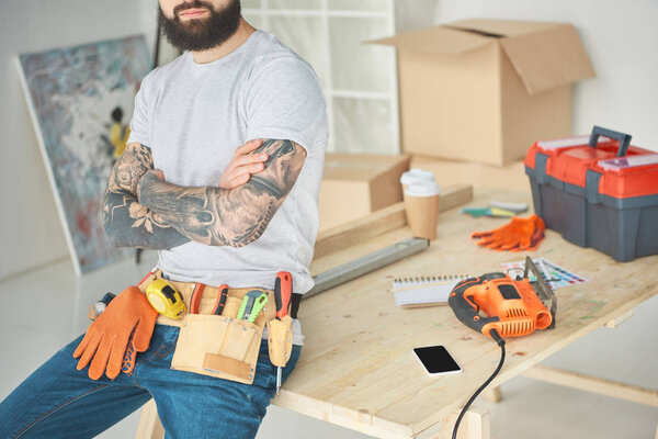 cropped shot of bearded tattooed man with tool belt sitting with crossed arms on wooden table