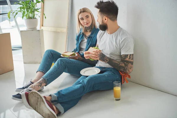 Young Tattooed Couple Eating Sandwiches Looking Each Other While Sitting — Free Stock Photo