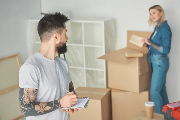 Bearded man taking notes and looking at girlfriend unpacking cardboard boxes in new house — Stock Image Bearded Man Taking Notes Looking Girlfriend Unpacking Cardboard Boxes New Stock Picture