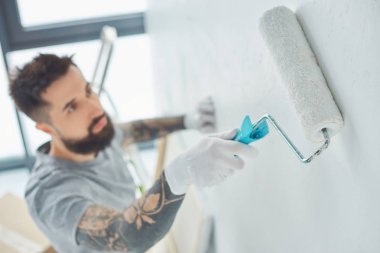 selective focus of tattooed repairman with roll paint brush painting wall in new apartment clipart