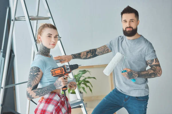 tattooed couple with screw gun and roll paint brush standing at ladder at new home
