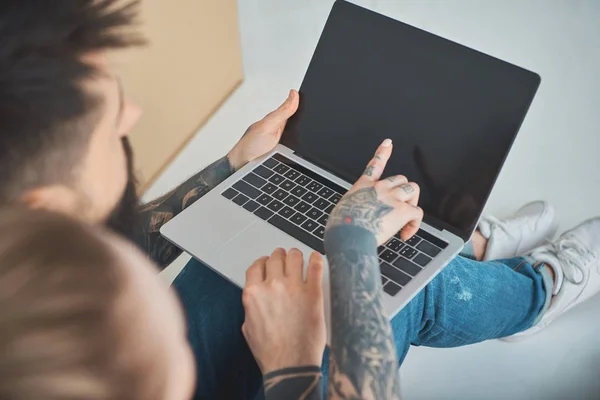 partial view of tattooed man pointing at blank laptop screen with girlfriend near by at new home