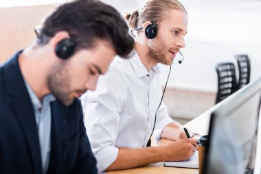 selective focus of male call center operators in headsets at workplace in office clipart
