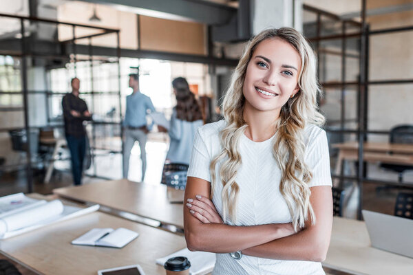 красивая молодая деловая женщина, стоящая со скрещенными руками и улыбающаяся в офисе перед камерой
