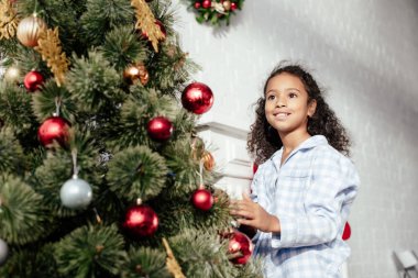 gülümseyen sevimli Afro-Amerikan çocuk baubles ile Noel ağacı süsleme ve uzak evde seyir pijama