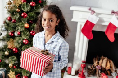 gülümseyen sevimli Afro-Amerikan çocuk Noel hediyesi ve seyir kameraya evde tutan pijama