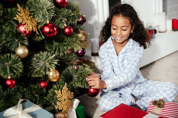 happy adorable african american child in pajamas decorating christmas tree with baubles at home