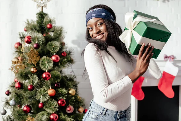 Счастливая Красивая Африканская Американка Держит Рождественский Подарок Дома Смотрит Камеру — Бесплатное стоковое фото