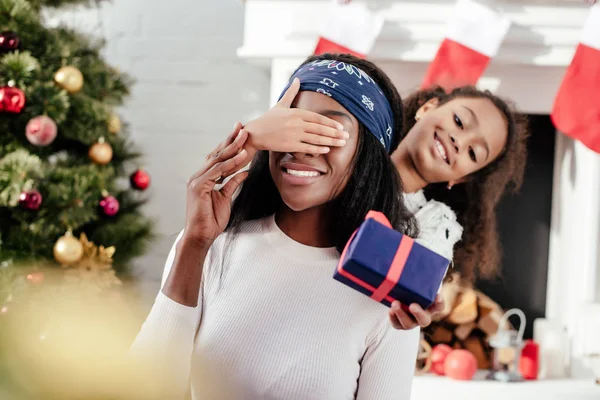 african american daughter gifting christmas present to mother and closing her eyes for surprise at home