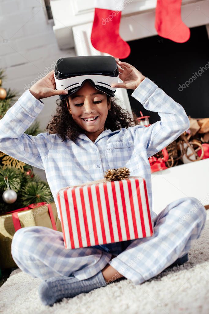 happy adorable african american child in pajamas and virtual reality headset looking at christmas gift at home