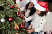 afro-amerikai anya és lánya a santa claus kalap díszítő karácsonyi fa együtt otthon
