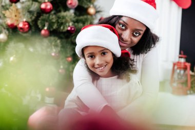 Afrika kökenli Amerikalı anne Noel Baba şapkalı bir şekilde dekore edilmiş oda evde küçük kızını sarılma