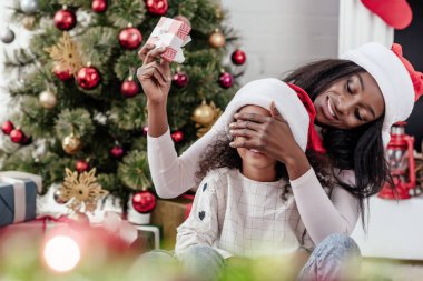 santa claus şapka şaşırtıcı kız Noel hediyesi ile Afrika kökenli Amerikalı anne evde gülümseyen
