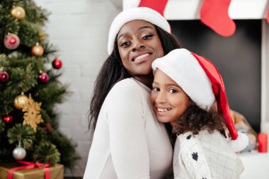 mutlu Afrika kökenli Amerikalı anne ve kızı, Noel Baba şapkaları dekore edilmiş oda evde portresi