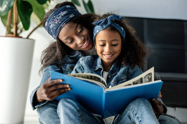 portrait of smiling african american mother and daughter looking at family photo album at home