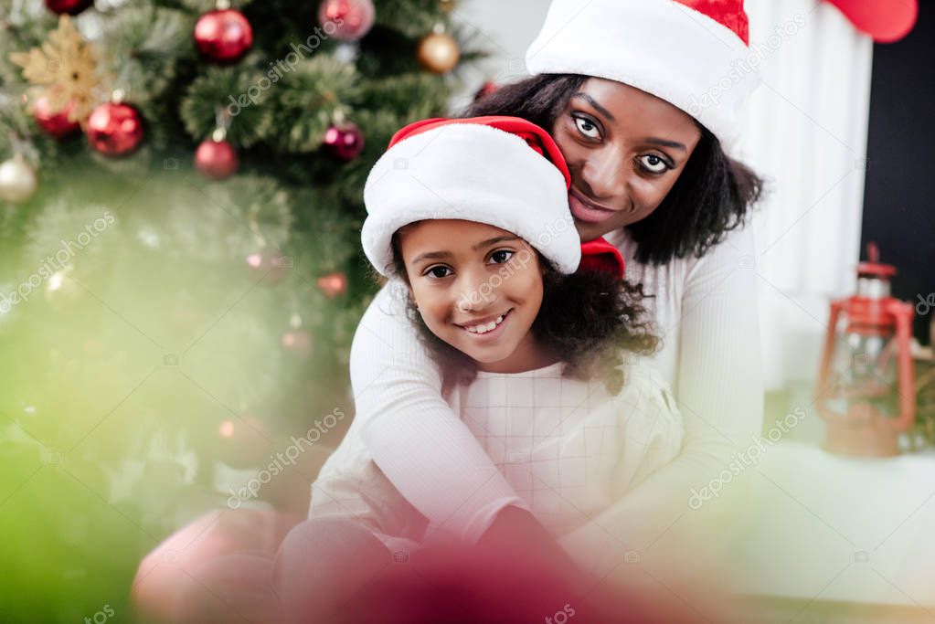 african american mother in santa claus hat hugging little daughter in decorated room at home