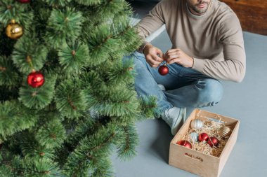 cropped image of man decorating christmas tree with baubles and sitting on floor at home clipart