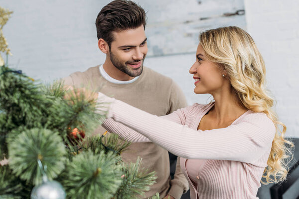 happy couple decorating christmas tree with baubles together at home and looking at each other