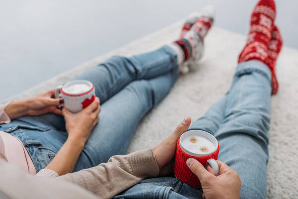 cropped image of couple holding cups of cappuccino and sitting on carpet at home