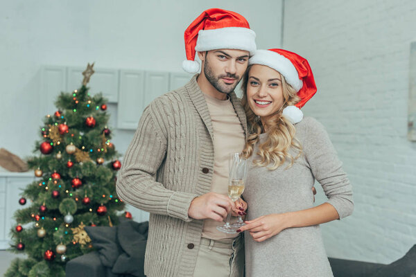 happy young couple with champagne glasses looking at camera in front of christmas tree at home