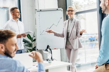 beyaz tahta duran ve Office erkek meslektaşları bakarak gülümseyen genç iş kadını