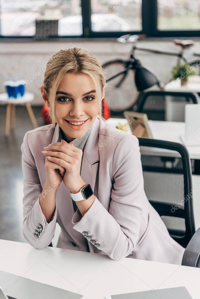 beautiful young businesswoman sitting at workplace and smiling at camera
