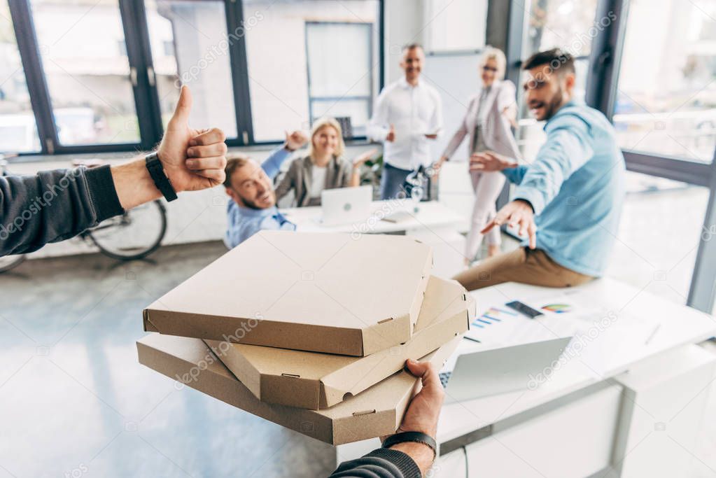 cropped shot of man holding pizza boxes and showing thumb up to colleagues in office