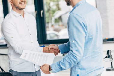 cropped shot of smiling businessmen holding contract and shaking hands in office clipart